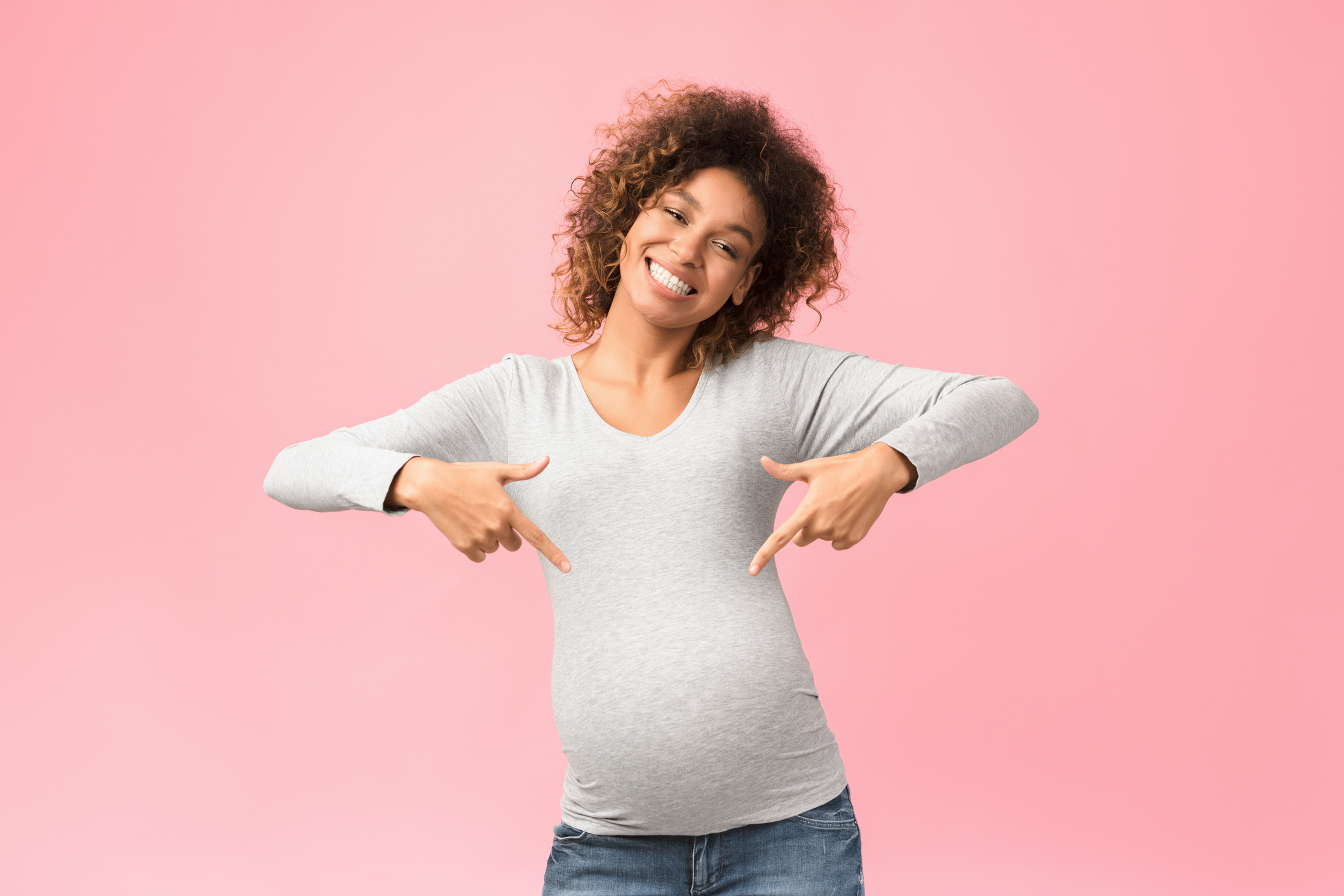 Look here. Positive afro expectant woman pointing fingers at her belly, pink background